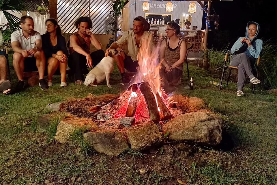 De noche, un grupo de huéspedes y un perro blanco se sientan junto a una fogata sobre el pasto; detrás brilla el pequeño Bar Bolívar.