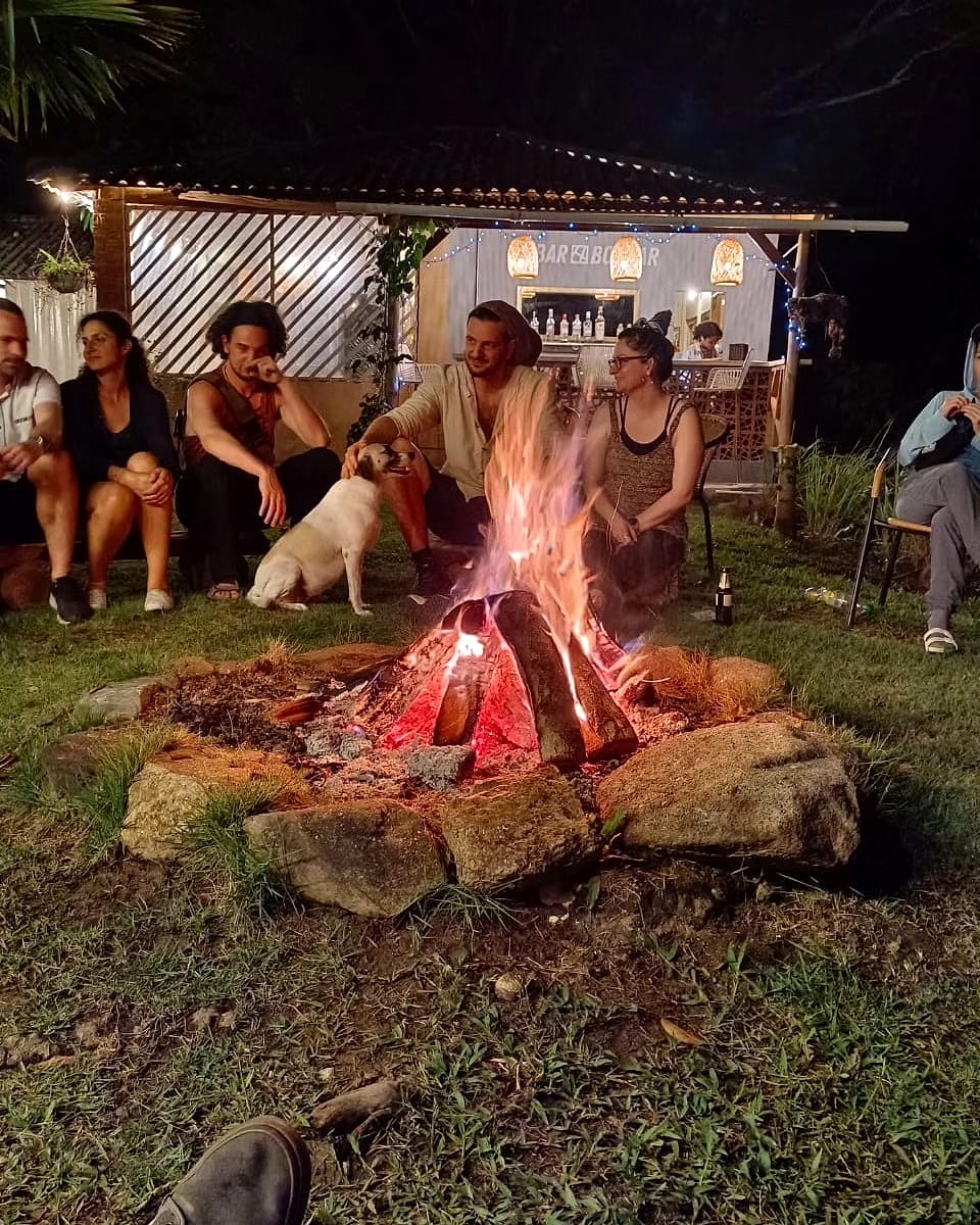 De noche, un grupo de huéspedes y un perro blanco se sientan junto a una fogata sobre el pasto; detrás brilla el pequeño Bar Bolívar.