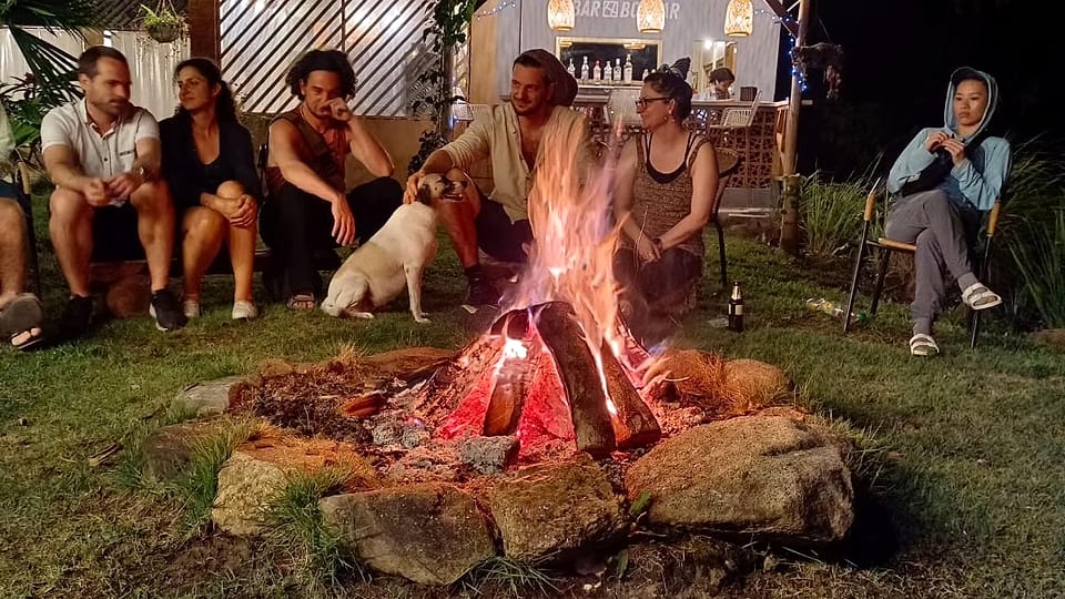 De noche, un grupo de huéspedes y un perro blanco se sientan junto a una fogata sobre el pasto; detrás brilla el pequeño Bar Bolívar.