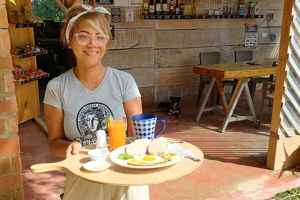 Una mujer del equipo, con camiseta de Finca Bolívar, sonríe sosteniendo una bandeja con huevos, pan, jugo de naranja y café; detrás, la barra y un perro echado.