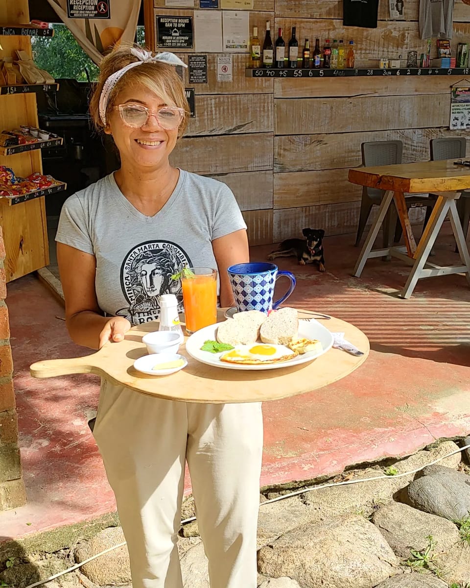 Una mujer del equipo, con camiseta de Finca Bolívar, sonríe sosteniendo una bandeja con huevos, pan, jugo de naranja y café; detrás, la barra y un perro echado.