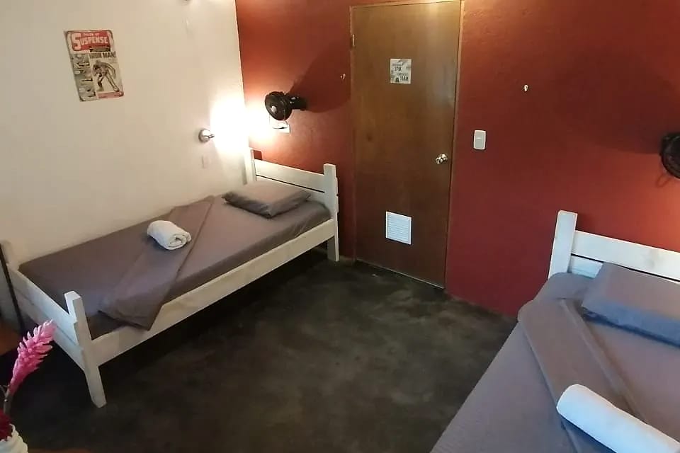 Habitación sencilla con pared roja, dos camas individuales con sábanas grises, ventilador de pared, lámparas de lectura y una puerta de madera.