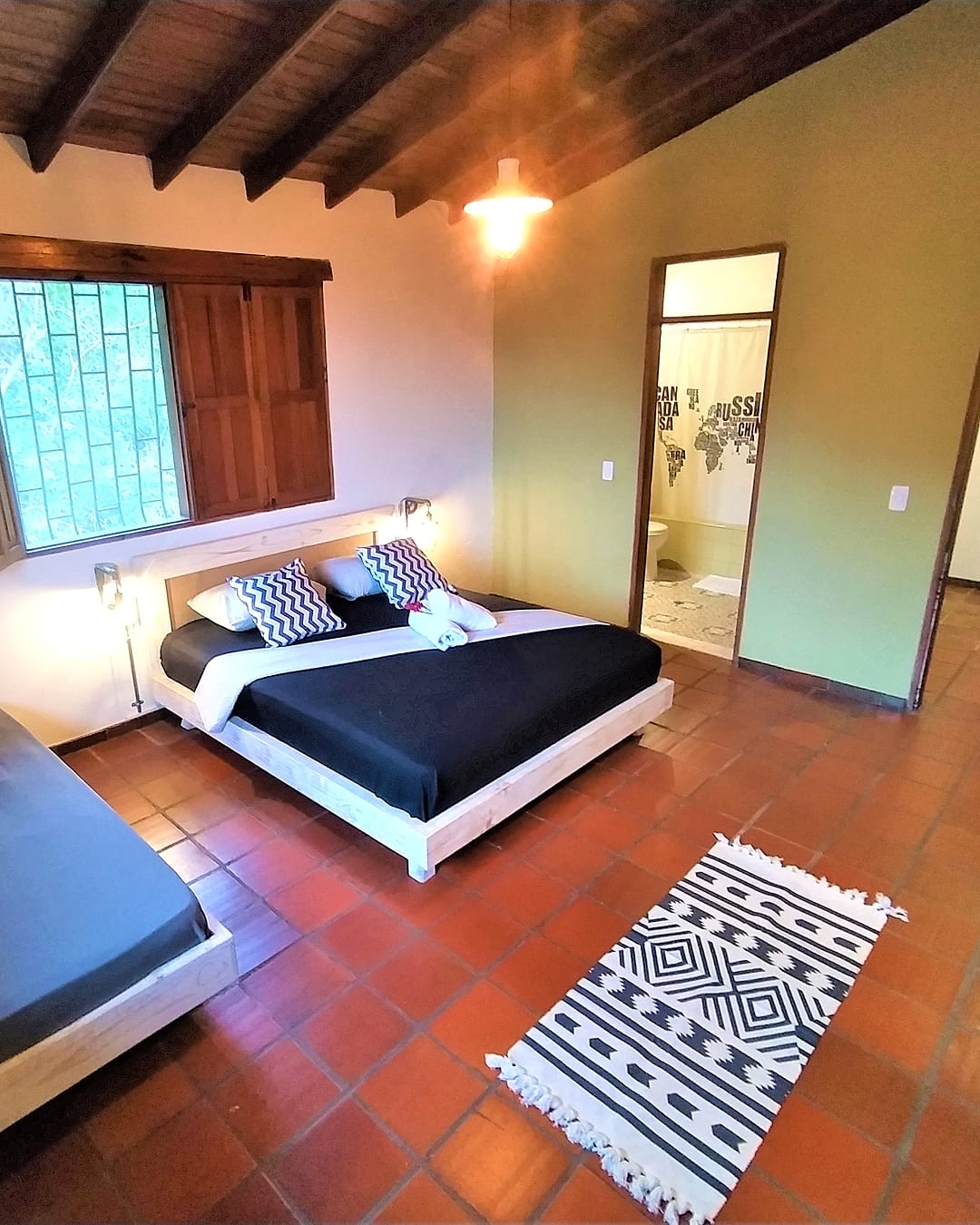 Habitación con cama doble de sábanas negras, un sofá cama, piso de baldosa de barro, techo de vigas de madera y ventanas con postigos; al fondo, la puerta del baño privado.