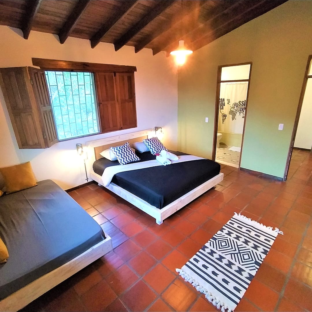 Habitación con cama doble de sábanas negras, un sofá cama, piso de baldosa de barro, techo de vigas de madera y ventanas con postigos; al fondo, la puerta del baño privado.