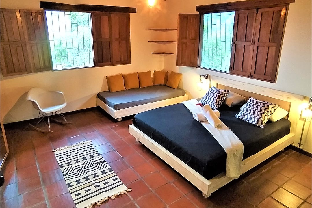 La misma habitación doble vista desde otro ángulo: el sofá cama con cojines amarillos bajo la ventana, estantes de madera y la cama doble.