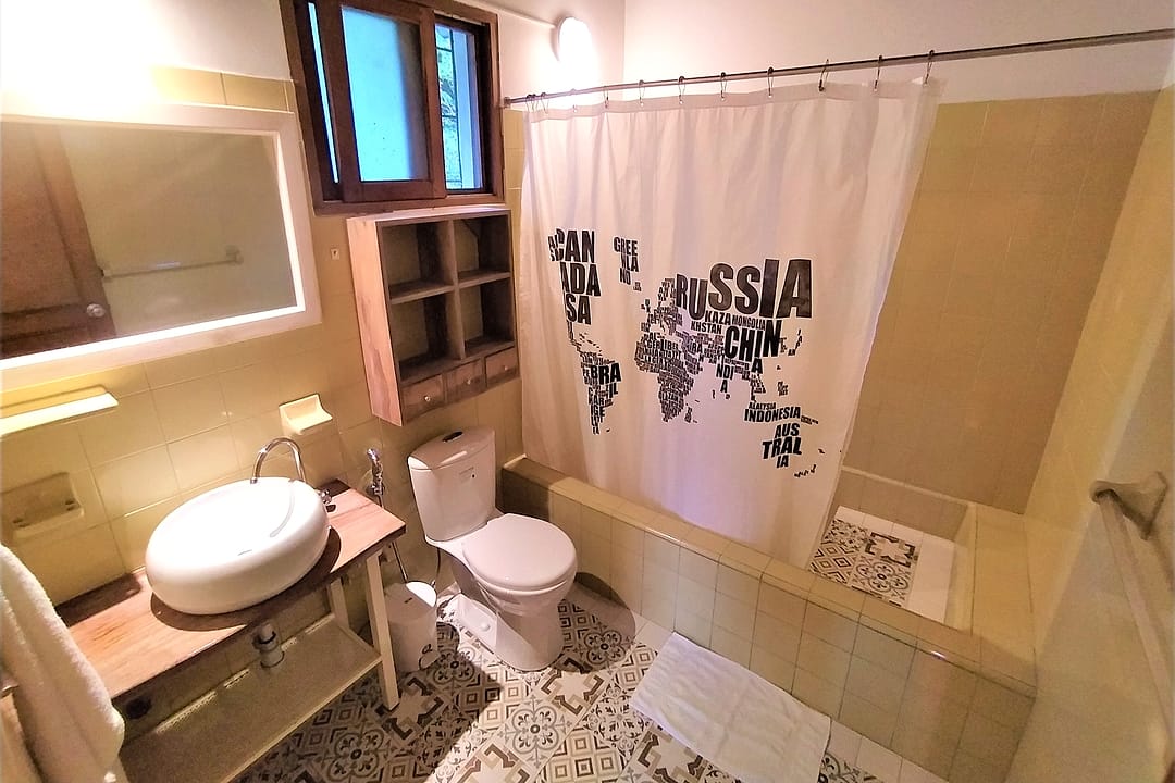 Baño privado con lavamanos de cerámica sobre repisa de madera, inodoro y una cortina de ducha estampada con un mapamundi.