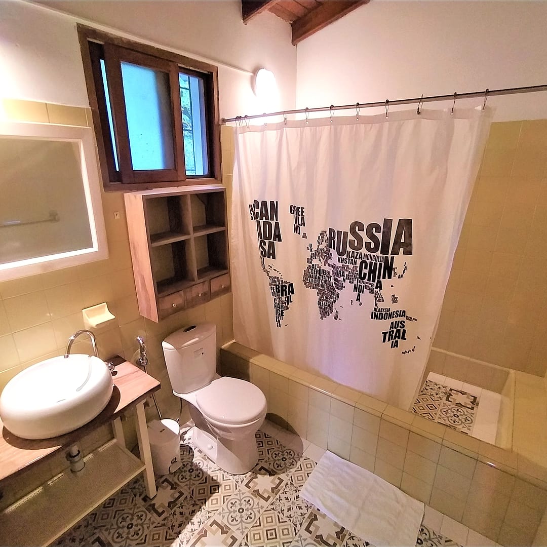 Baño privado con lavamanos de cerámica sobre repisa de madera, inodoro y una cortina de ducha estampada con un mapamundi.
