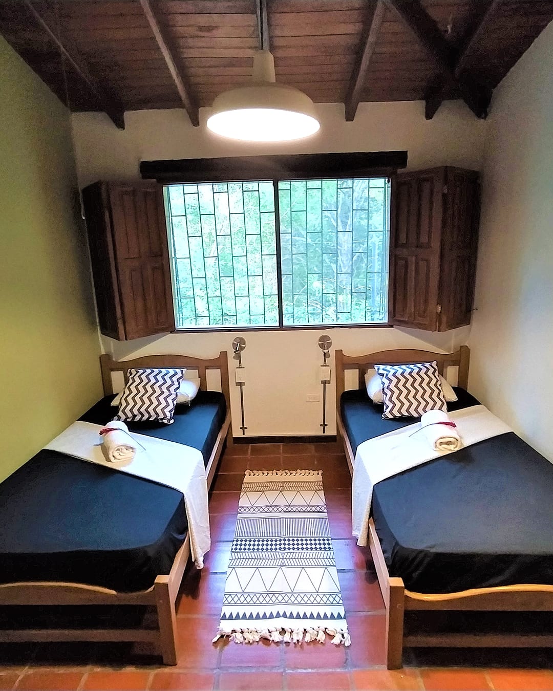 Habitación con dos camas sencillas de sábanas negras, una ventana con reja y postigos de madera, lámpara de techo y tapete tejido entre las camas.