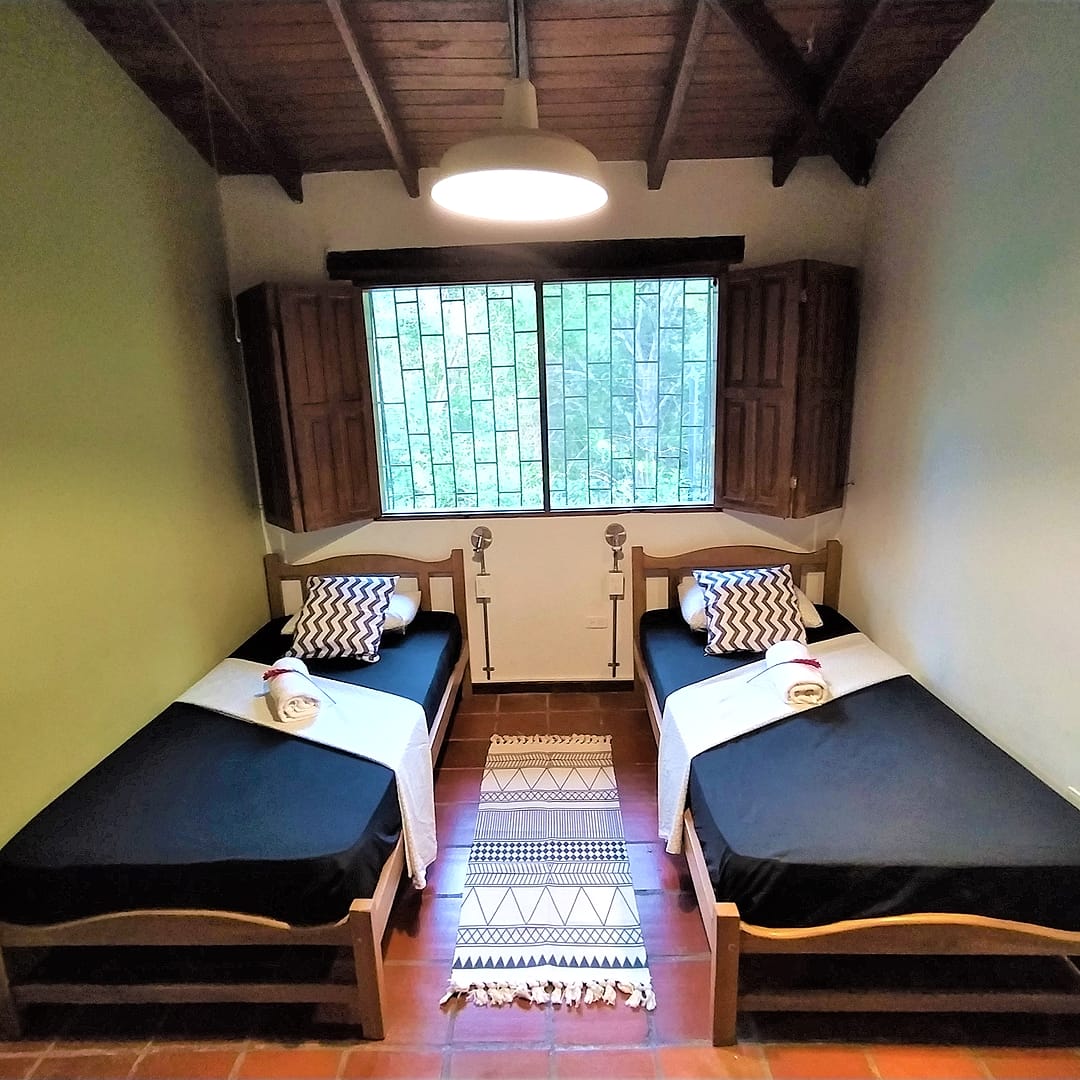 Habitación con dos camas sencillas de sábanas negras, una ventana con reja y postigos de madera, lámpara de techo y tapete tejido entre las camas.