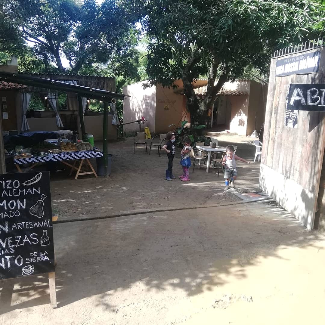 Patio de tierra bajo árboles grandes, con un puesto de charcutería, pizarras escritas a mano, mesas y niños jugando al fondo.