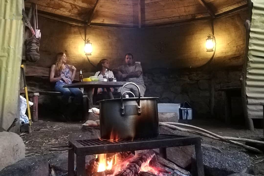 Bajo el techo de palma de una choza junto al río, una olla de sancocho hierve sobre la leña mientras unos huéspedes conversan a la luz de faroles.