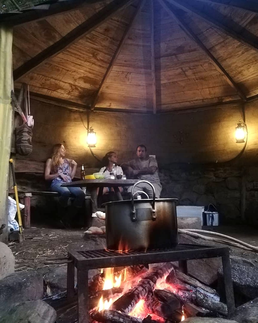 Bajo el techo de palma de una choza junto al río, una olla de sancocho hierve sobre la leña mientras unos huéspedes conversan a la luz de faroles.