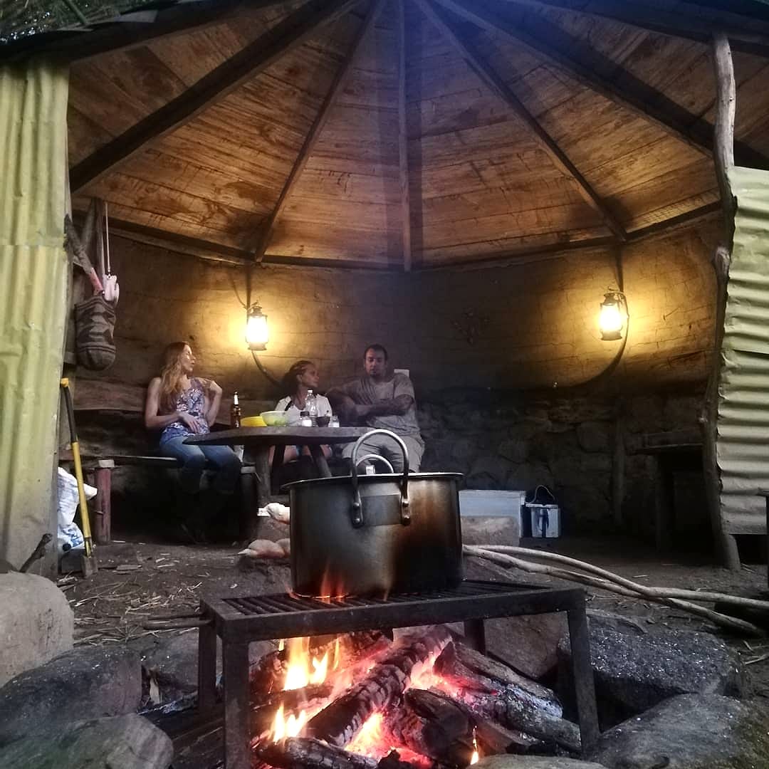 Bajo el techo de palma de una choza junto al río, una olla de sancocho hierve sobre la leña mientras unos huéspedes conversan a la luz de faroles.