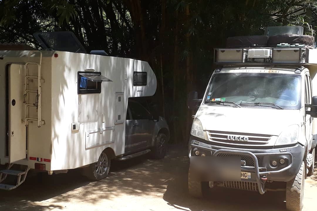 Una autocaravana blanca y un camión 4x4 estacionados a la sombra de bambúes en la finca.