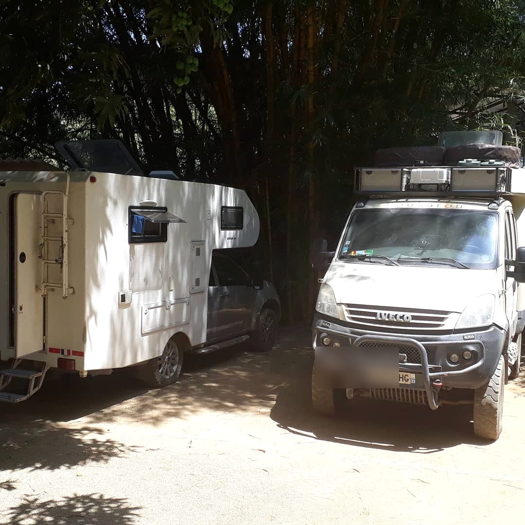 Una autocaravana blanca y un camión 4x4 estacionados a la sombra de bambúes en la finca.