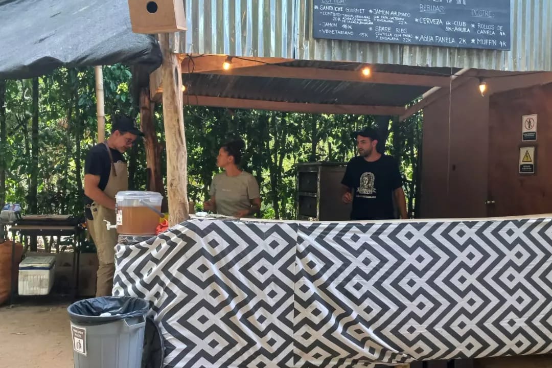 Un puesto de comida con techo de zinc, pizarra con el menú y barra cubierta con tela geométrica, atendido por dos personas.