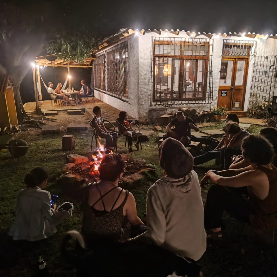 Una casita blanca con luces colgantes y ventanas encendidas; delante, huéspedes conversan sentados en la oscuridad.