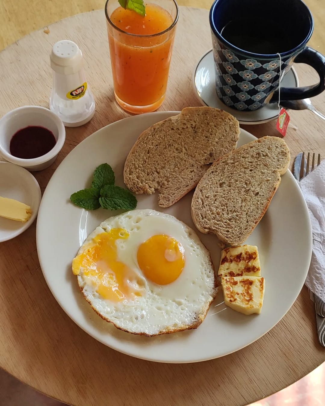 Plato de desayuno con huevo frito, pan casero, queso a la plancha, mantequilla y mermelada, con jugo de naranja y café en taza azul.