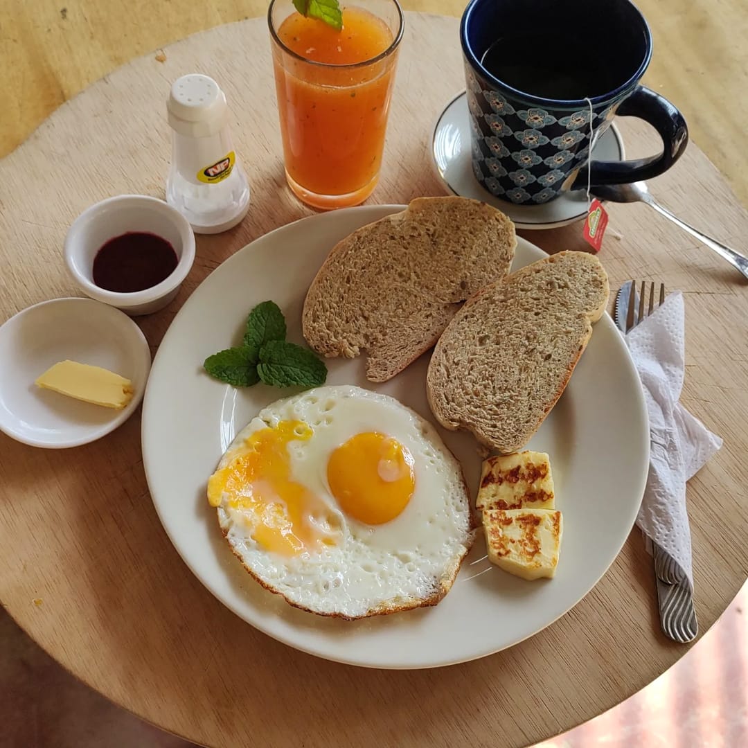 Plato de desayuno con huevo frito, pan casero, queso a la plancha, mantequilla y mermelada, con jugo de naranja y café en taza azul.