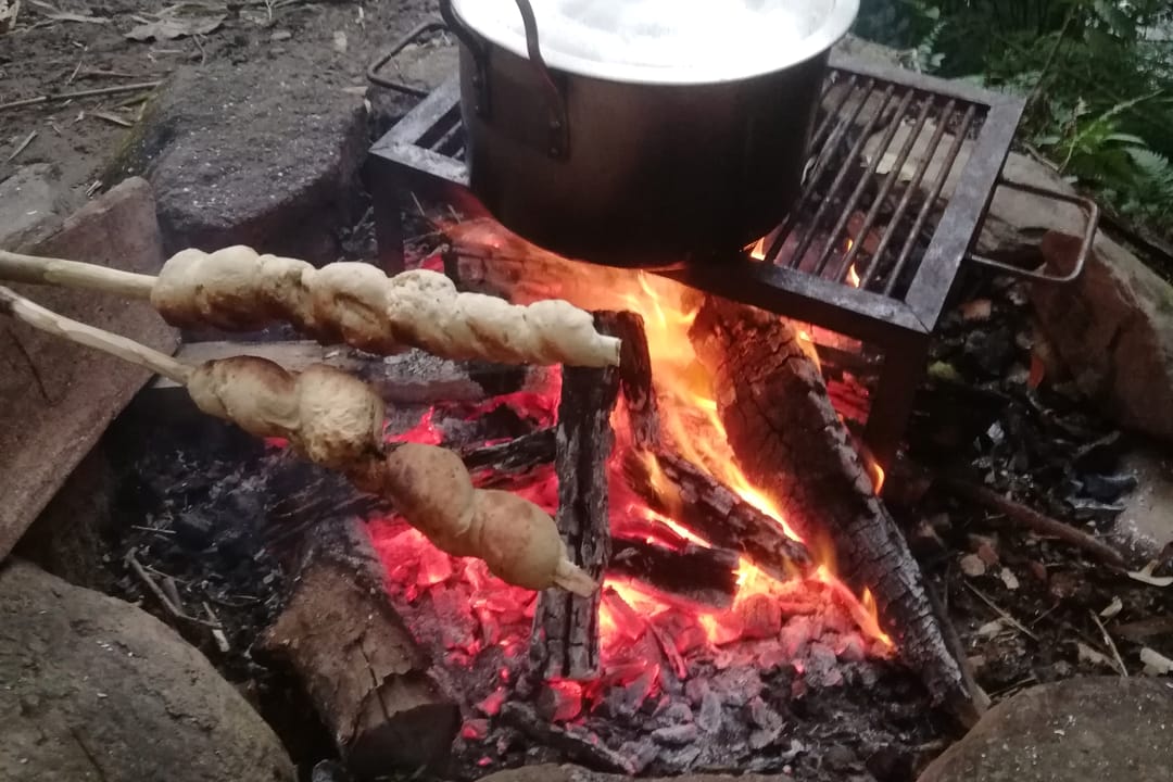 Masa de pan enrollada en palos asándose junto a una olla sobre las brasas.