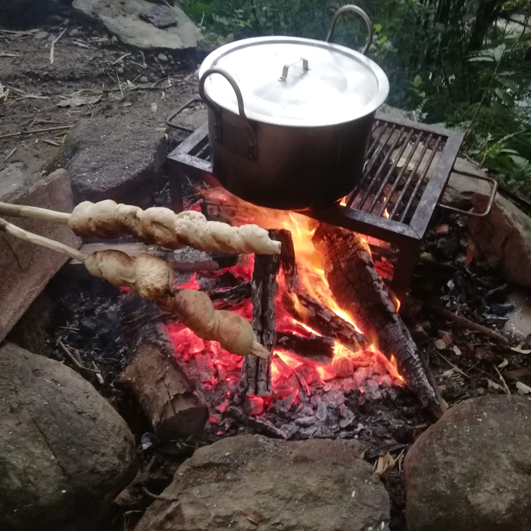 Masa de pan enrollada en palos asándose junto a una olla sobre las brasas.
