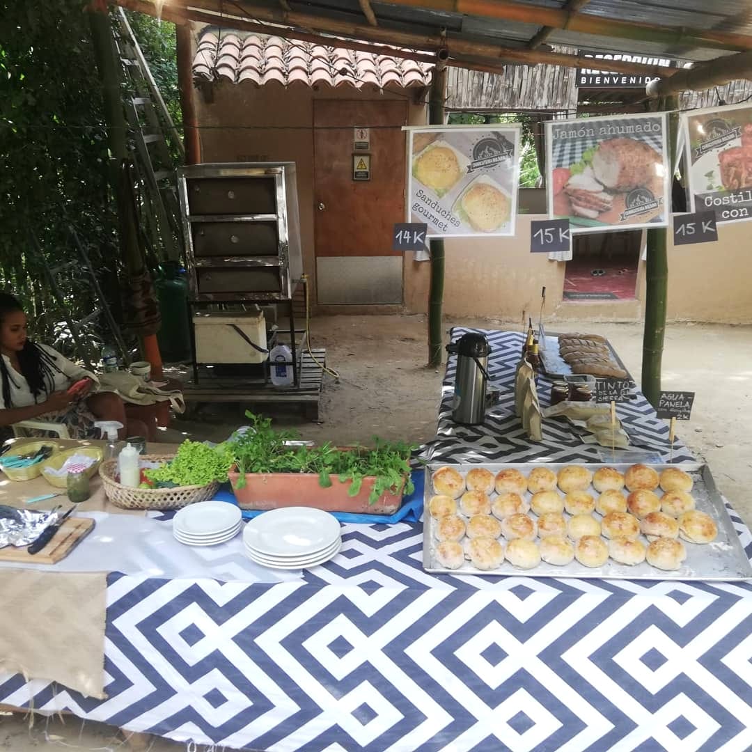 Mesa con mantel geométrico blanco y negro llena de pan, platos y embutidos bajo un toldo, con carteles de comida colgados.