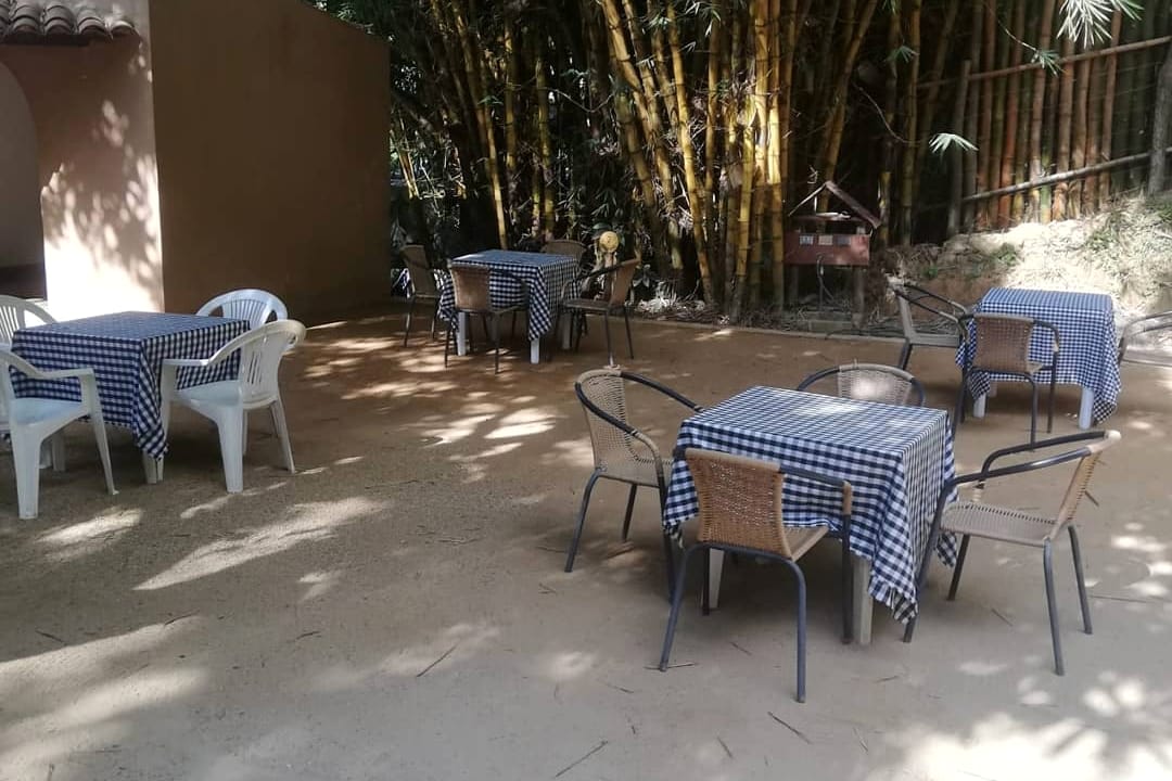 Patio de tierra con mesas de mantel a cuadros azules y sillas plásticas, rodeado de guaduales y casas bajas.