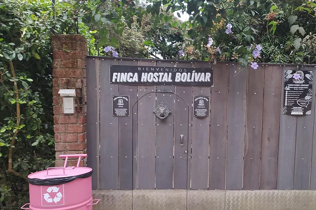 Portón de madera gris con el letrero “Finca Hostal Bolívar”, entre pilares de ladrillo y bajo flores moradas, al borde de la calle de concreto.