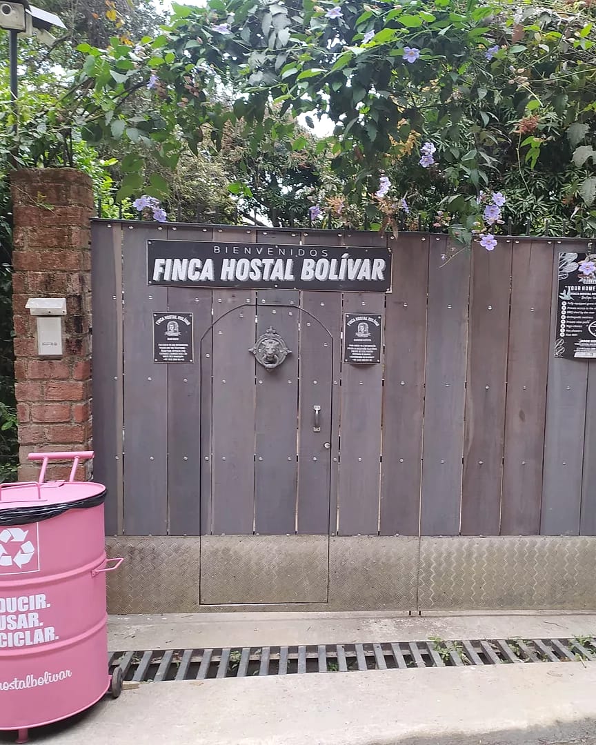 Portón de madera gris con el letrero “Finca Hostal Bolívar”, entre pilares de ladrillo y bajo flores moradas, al borde de la calle de concreto.