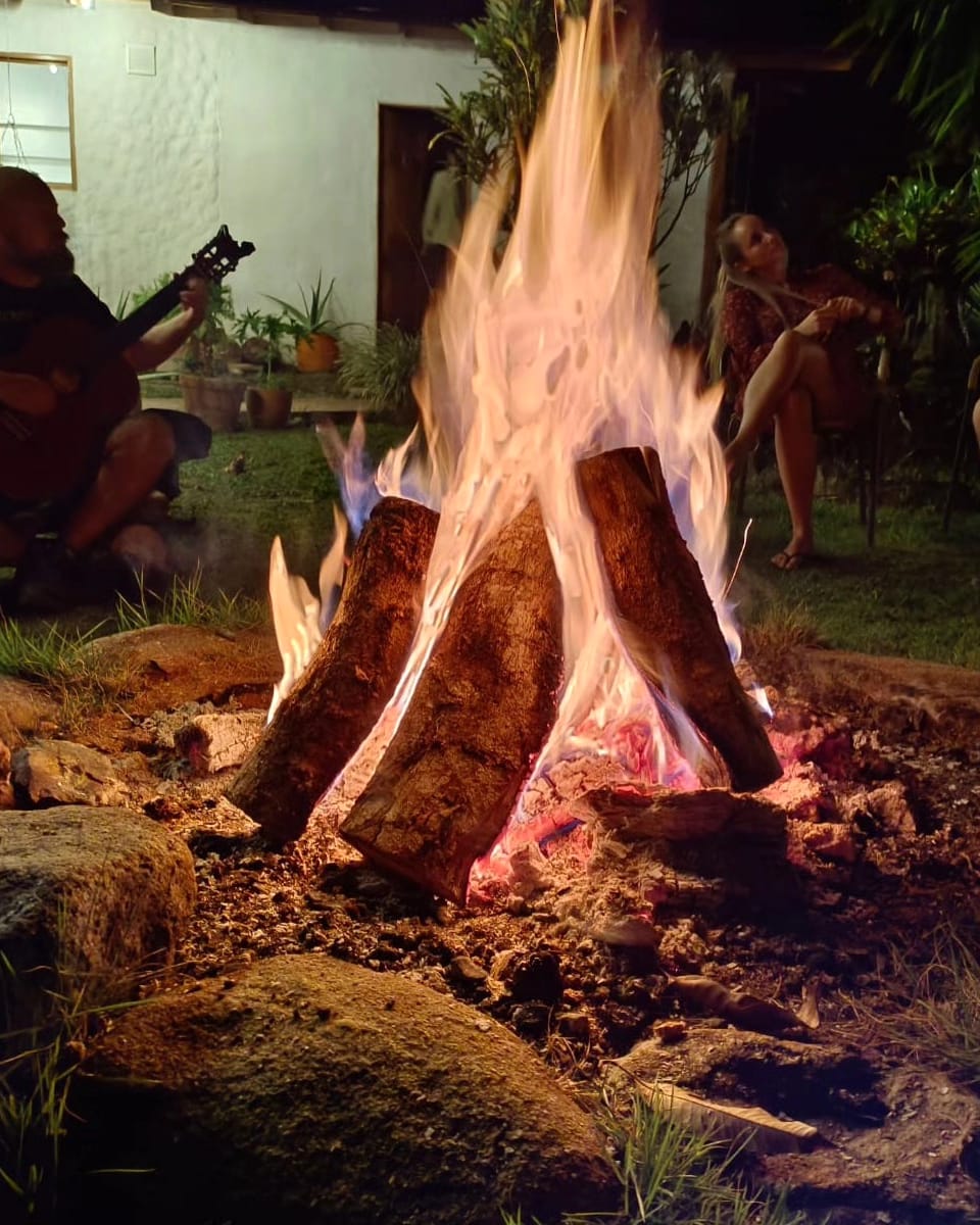 Primer plano de una fogata alta; a un lado alguien toca la guitarra y otros escuchan en la penumbra.