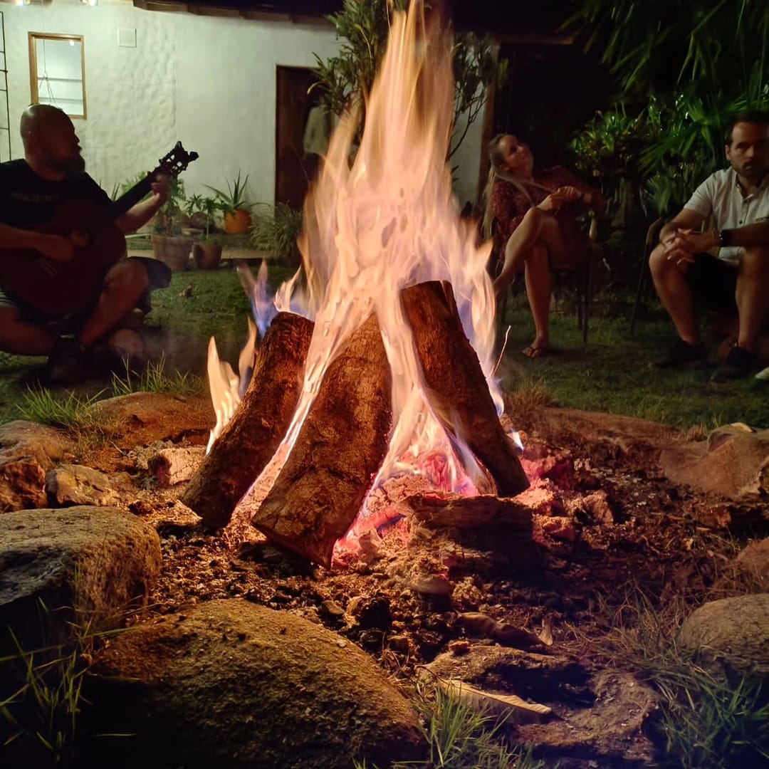 Primer plano de una fogata alta; a un lado alguien toca la guitarra y otros escuchan en la penumbra.