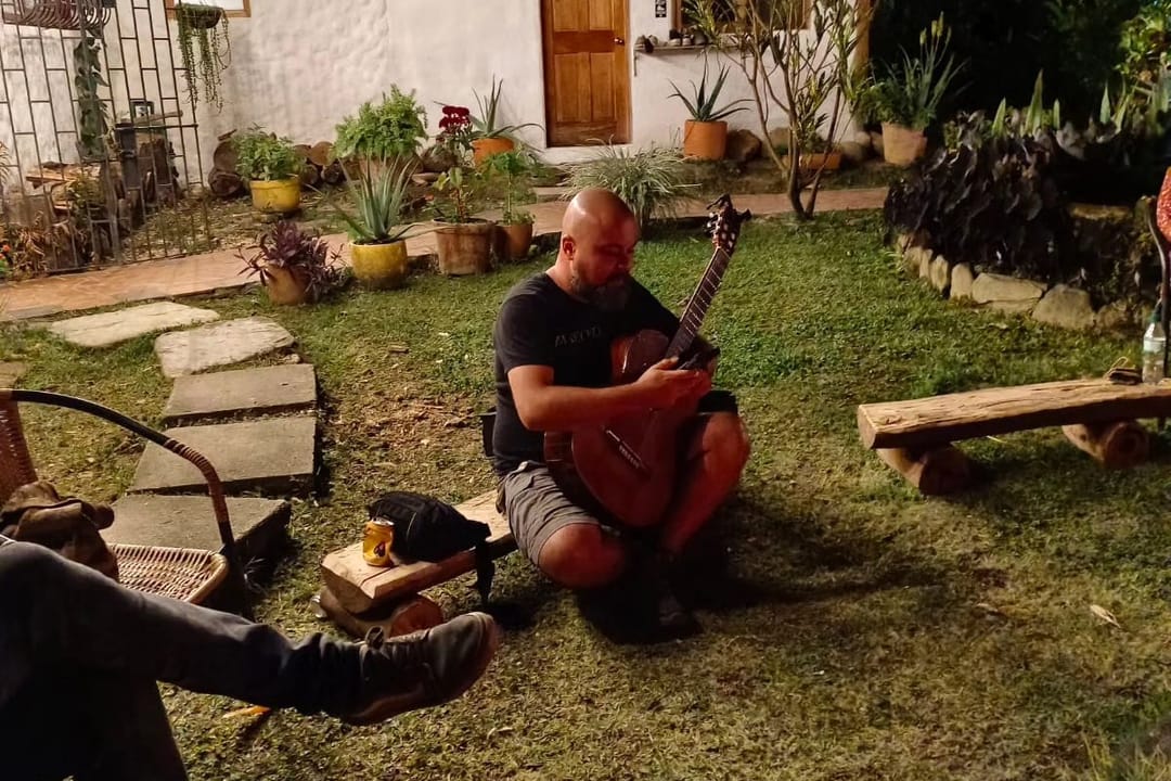 Un hombre toca la guitarra sentado en el pasto frente a una casa blanca con puerta de madera, macetas y un camino de lajas.