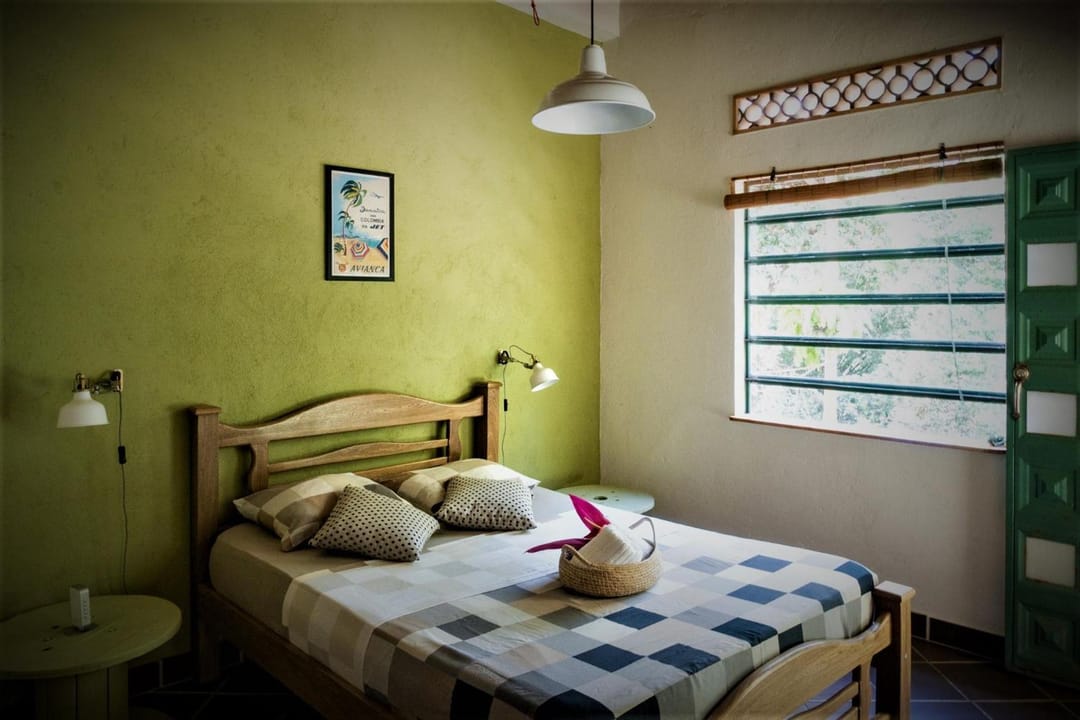 Habitación doble de pared verde: cama de madera, colcha de cuadros, ventana grande y lámpara colgante.