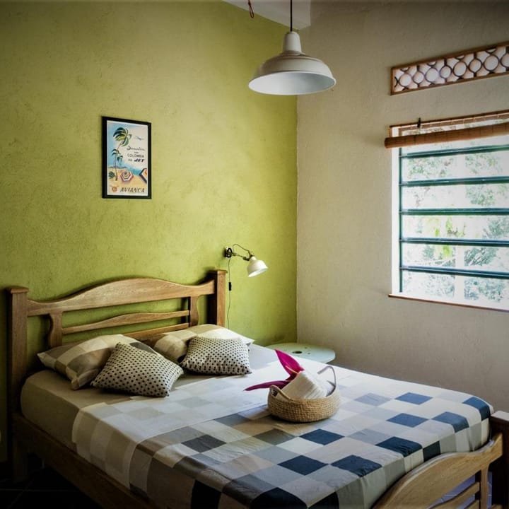 Habitación doble de pared verde: cama de madera, colcha de cuadros, ventana grande y lámpara colgante.