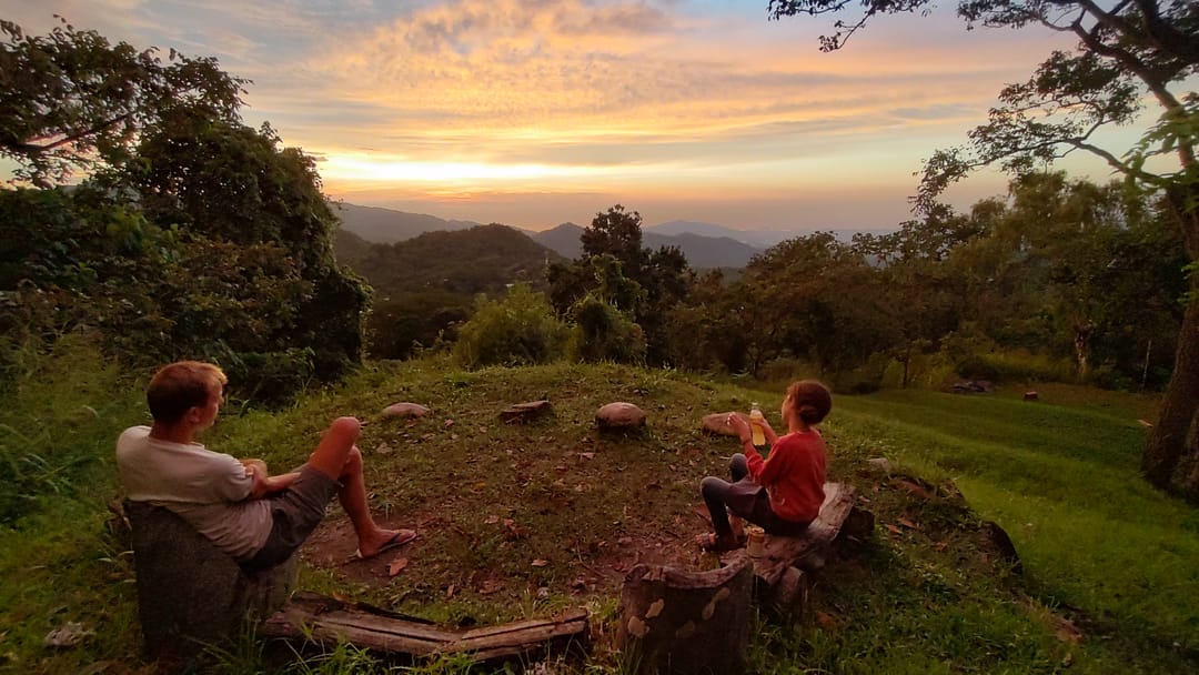 Dos personas sentadas en el pasto del mirador miran el atardecer sobre las lomas.