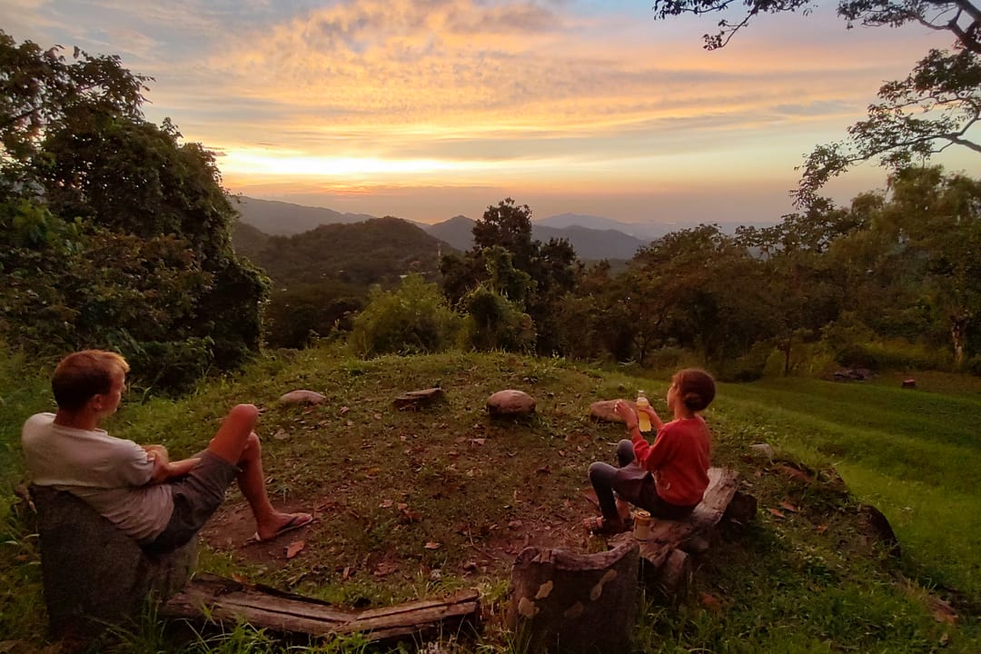 Dos personas sentadas en el pasto del mirador miran el atardecer sobre las lomas.