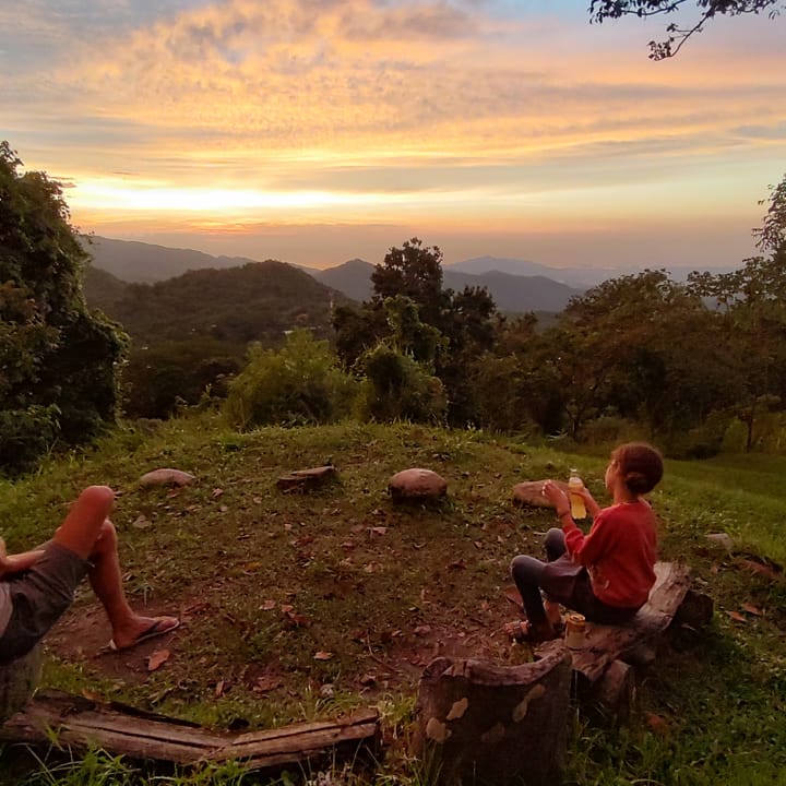 Dos personas sentadas en el pasto del mirador miran el atardecer sobre las lomas.