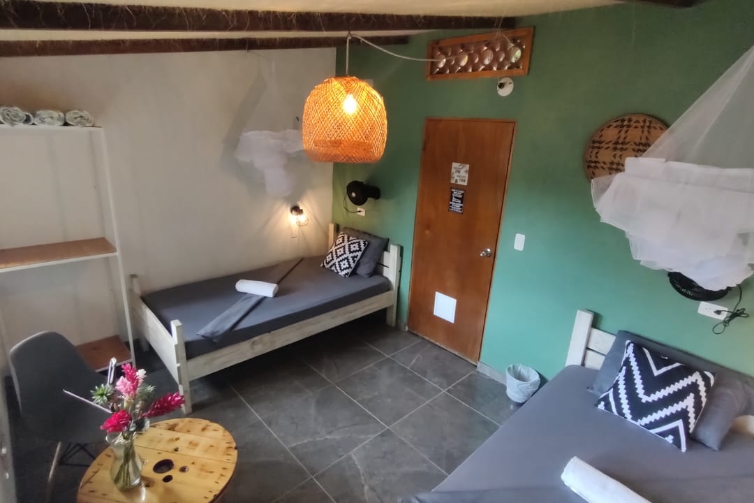 La twin de Casa Mango con pared verde, toldillo, lámpara tejida naranja y puerta de madera.