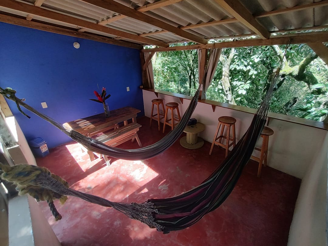 Zona común techada con hamacas, pared azul, piso rojo y mesa de madera, abierta a la selva.