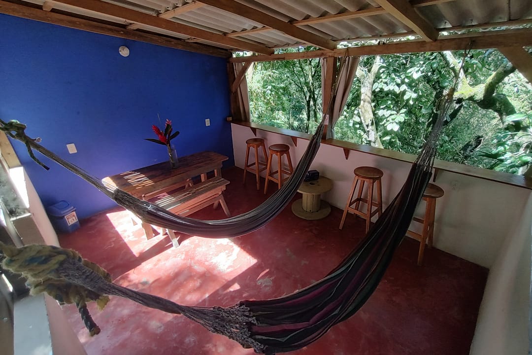 Zona común techada con hamacas, pared azul, piso rojo y mesa de madera, abierta a la selva.