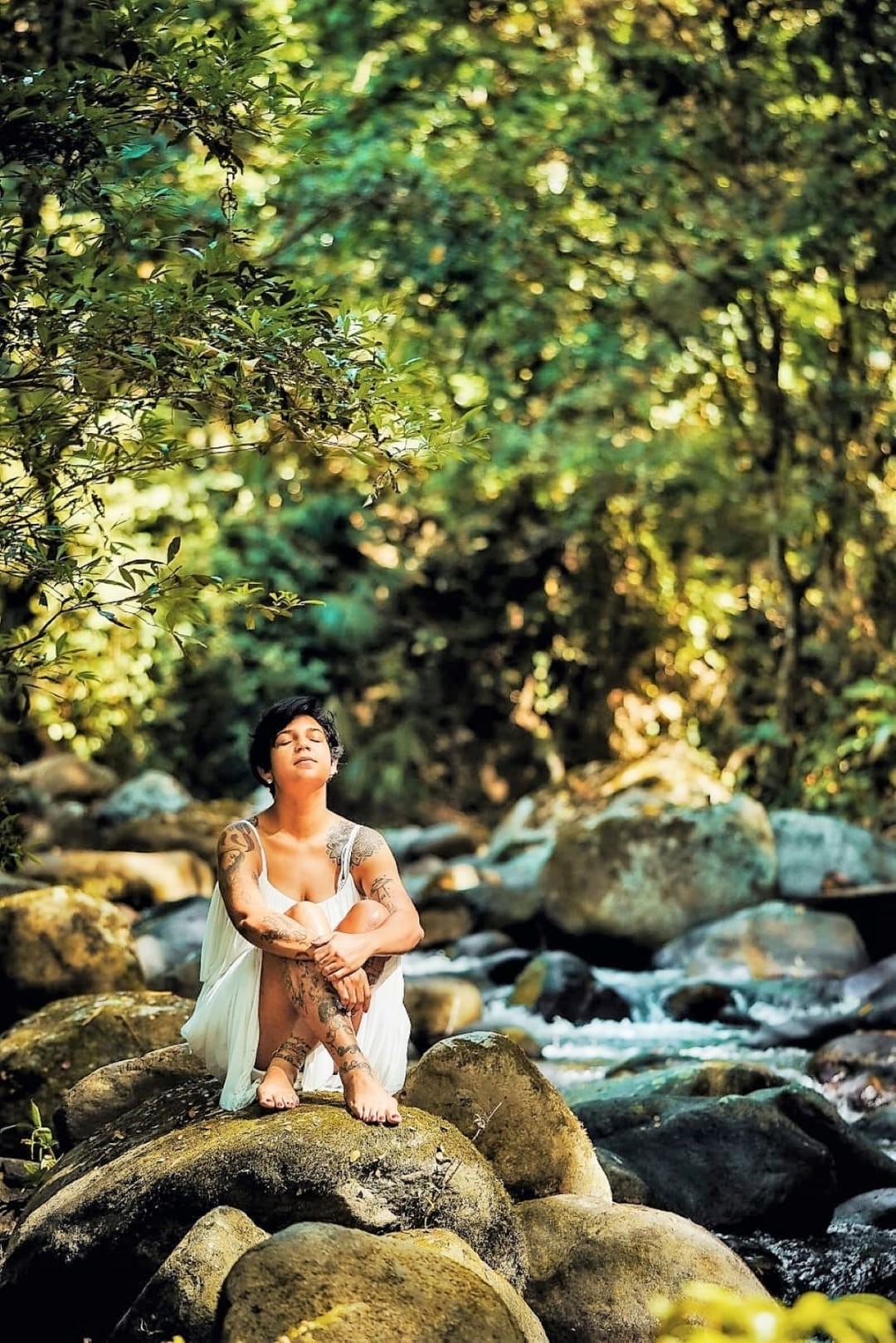 Una mujer sentada sobre las piedras del río, con luz entre los árboles.