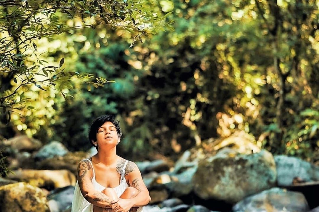 Una mujer sentada sobre las piedras del río, con luz entre los árboles.