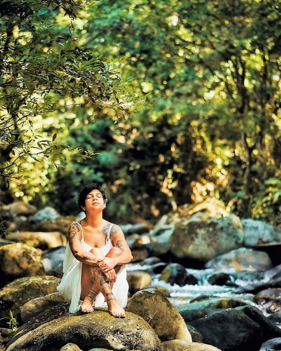 Una mujer sentada sobre las piedras del río, con luz entre los árboles.