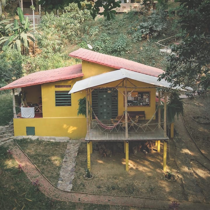 La casa amarilla sobre pilotes, con techo rosado, vista desde arriba entre la vegetación.