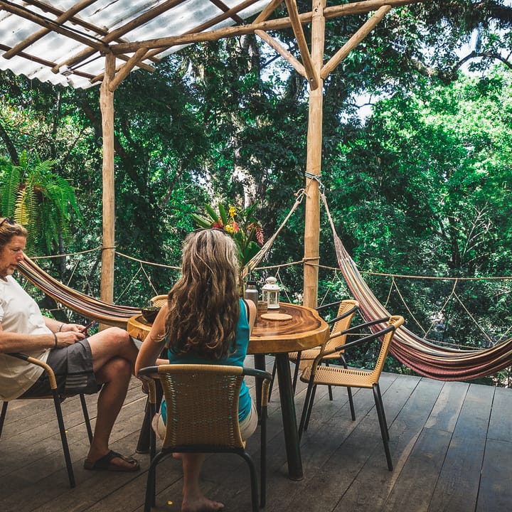 Dos personas conversan en la terraza de madera, con hamacas y selva alrededor.