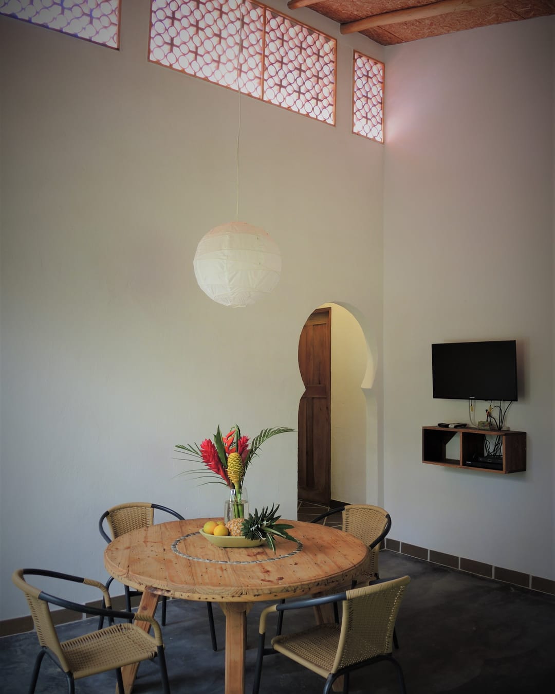 Comedor con mesa redonda, flores de heliconia, televisor y una puerta en arco.