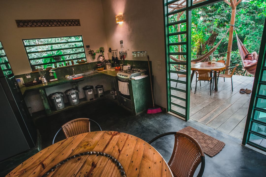 Cocina con ventanas verdes, mesa redonda de madera y la puerta abierta hacia la terraza de hamacas.