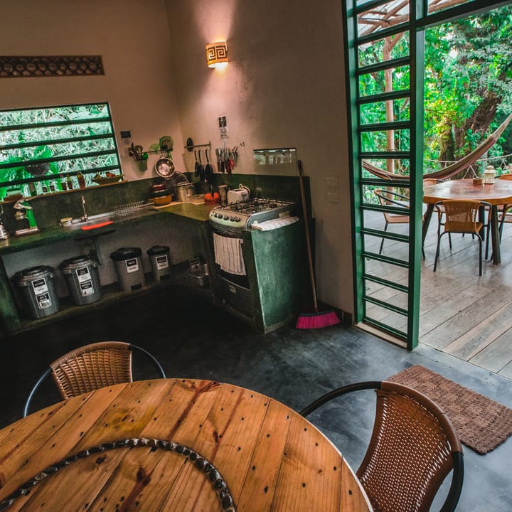 Cocina con ventanas verdes, mesa redonda de madera y la puerta abierta hacia la terraza de hamacas.