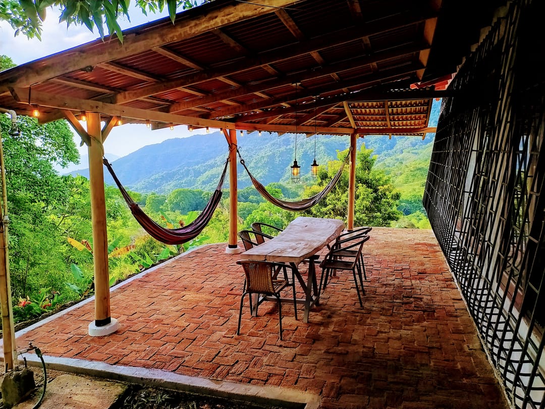 Terraza techada con piso de ladrillo, dos hamacas, mesa larga y el valle verde al fondo.
