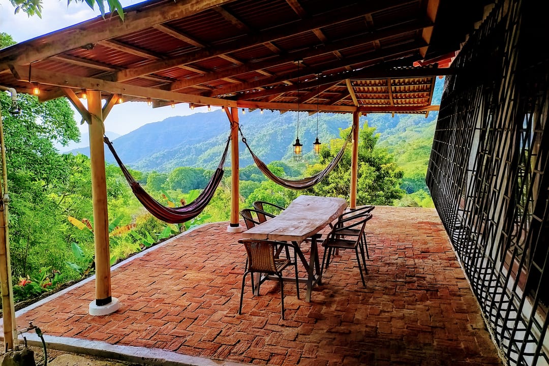 Terraza techada con piso de ladrillo, dos hamacas, mesa larga y el valle verde al fondo.